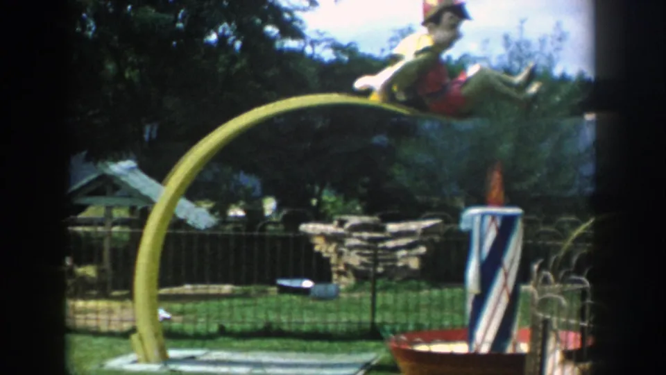 1954 Wisconsin Dells frame with a yellow arched ride, clown figure, striped post, low fencing, stone wall, and trees behind it.