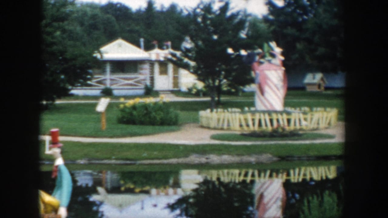 Lakefront amusement area with a white building, giant striped figure, lawns, and water reflection.