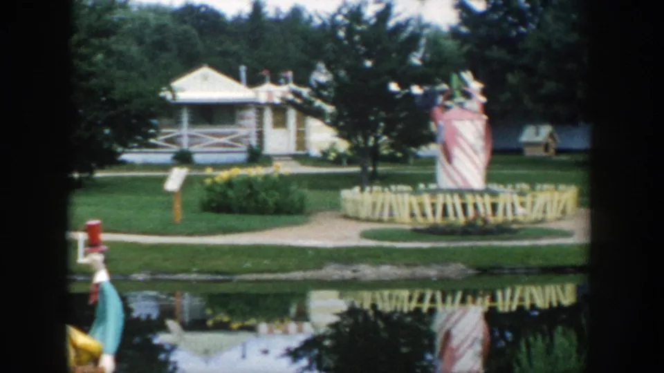1954 Wisconsin Dells 8mm frame showing a landscaped lakefront attraction with a white building, oversized striped figure, paths, trees, and reflections.