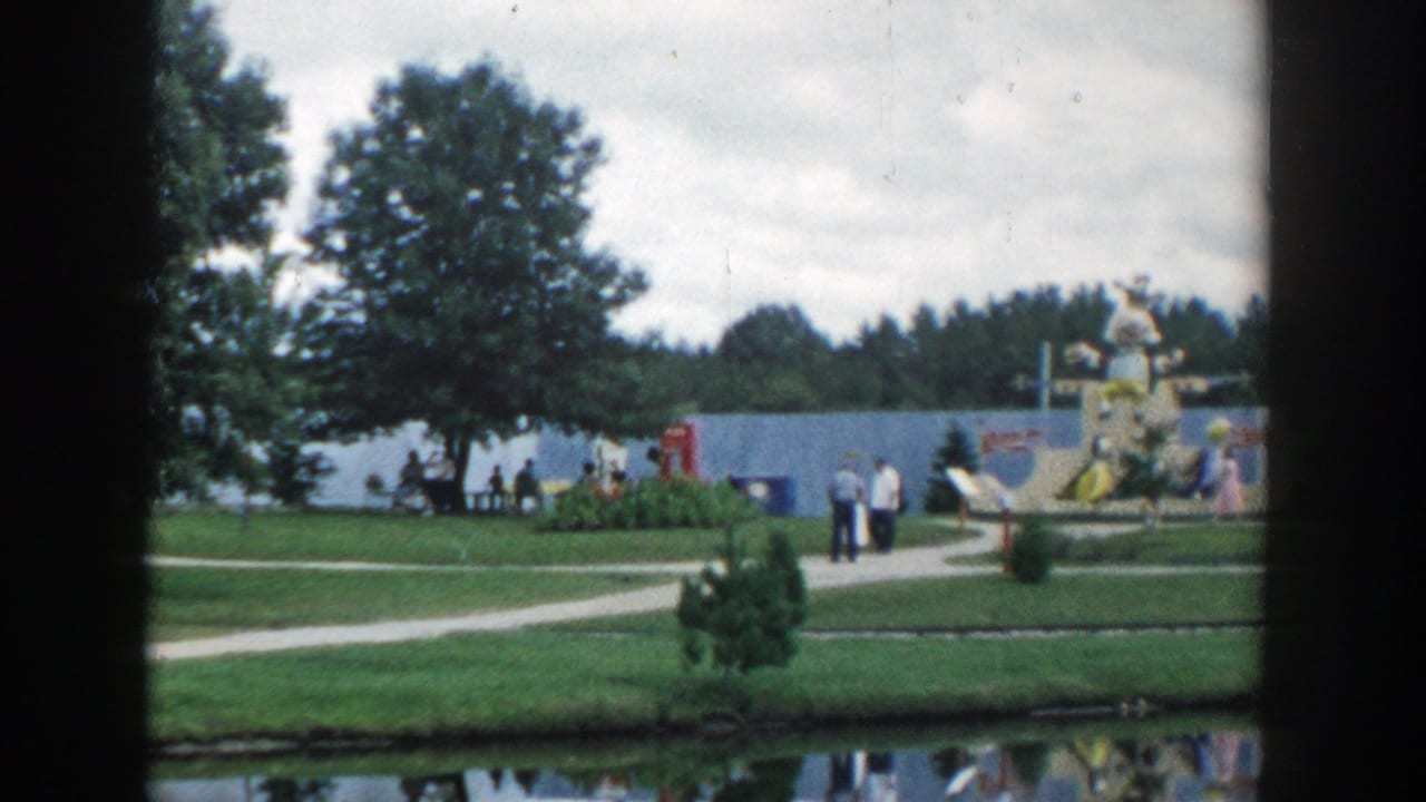 Park paths near a lake with visitors, trees, and colorful amusement figures in the distance.