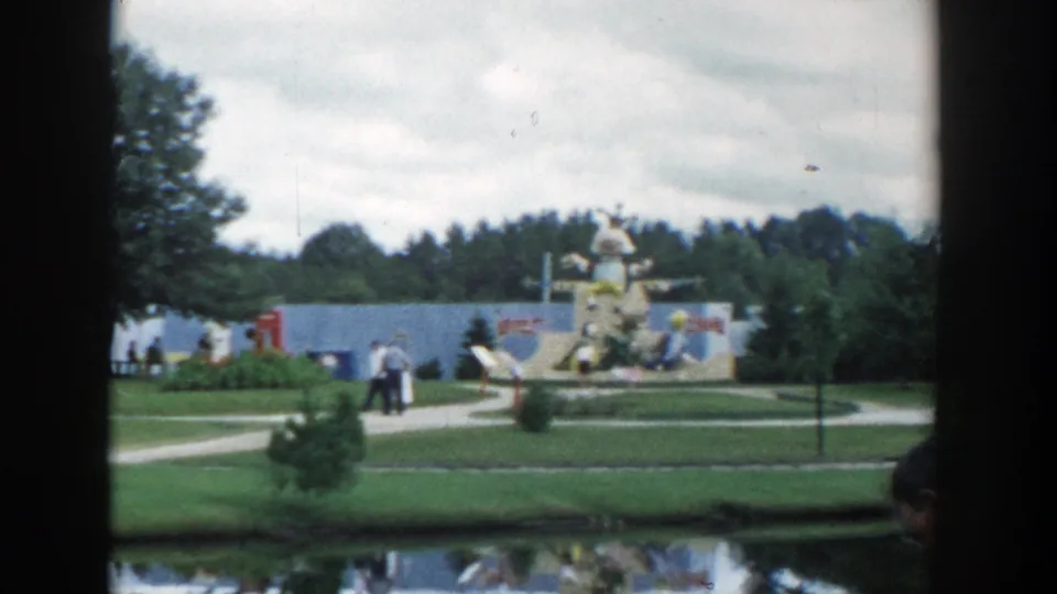 1954 Wisconsin Dells 8mm frame with visitors on a path, a small tree, lawns, and large decorative amusement figures beyond a reflective water edge.