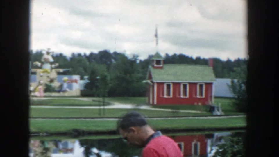 1954 Wisconsin Dells frame showing a red schoolhouse-style building, lawns, paths, reflective water, and an adult man partly entering the foreground.