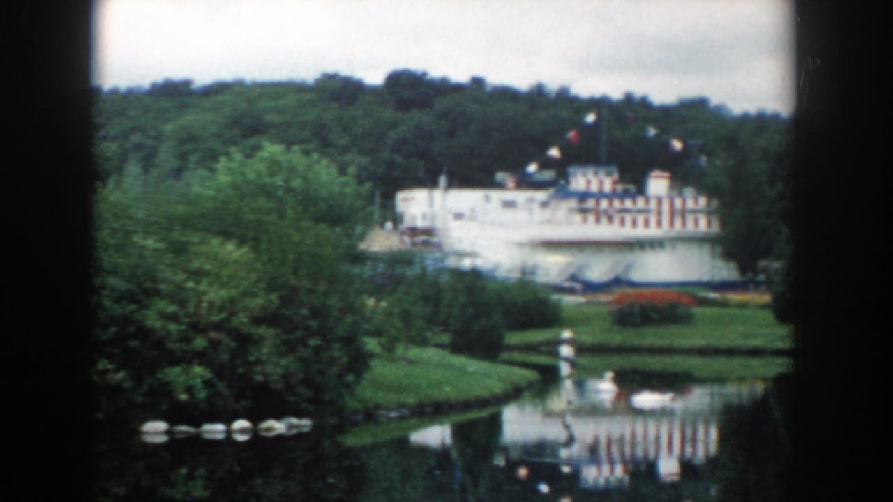 1954 Wisconsin Dells park view with a pond, dense trees, and a white building with red and blue striping beyond the water.