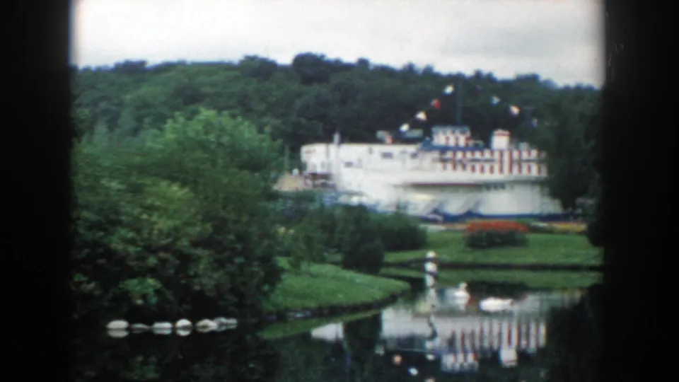 1954 Wisconsin Dells park view with a pond, dense trees, and a white building with red and blue striping beyond the water.