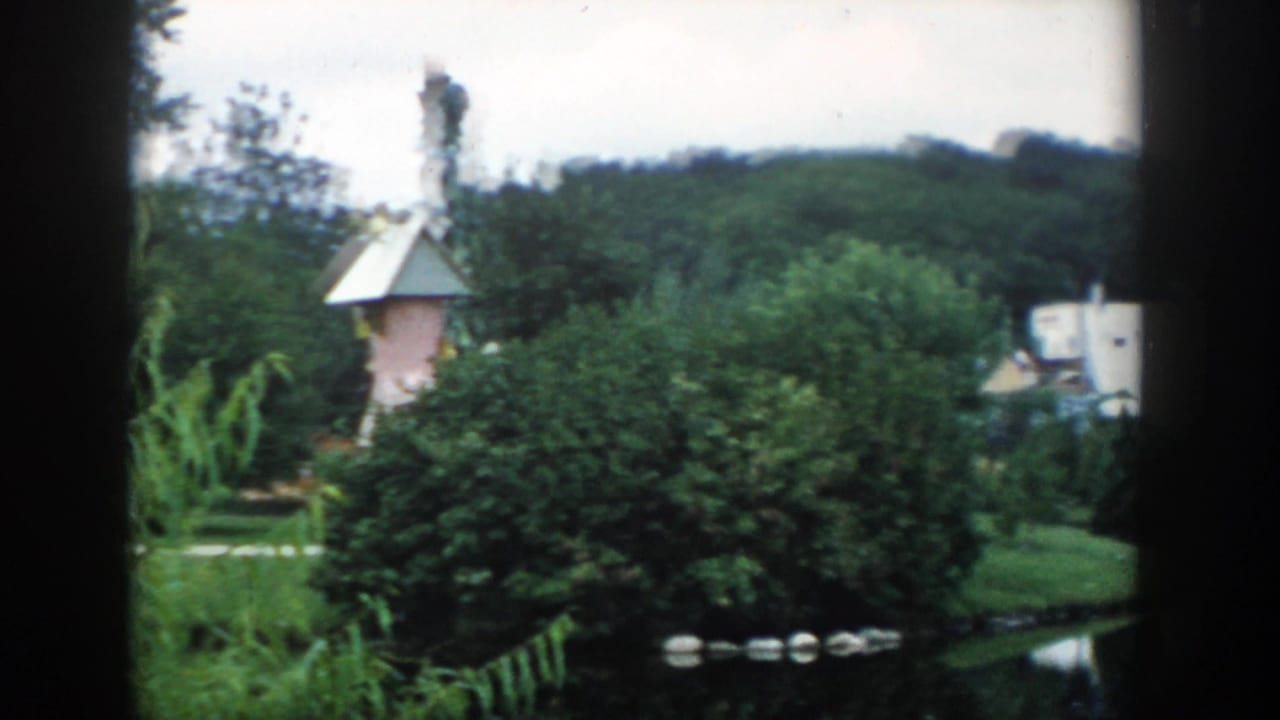 1954 Wisconsin Dells garden scene showing shrubs, lawn, water, and a small pink tower-like structure near the landscaped grounds.