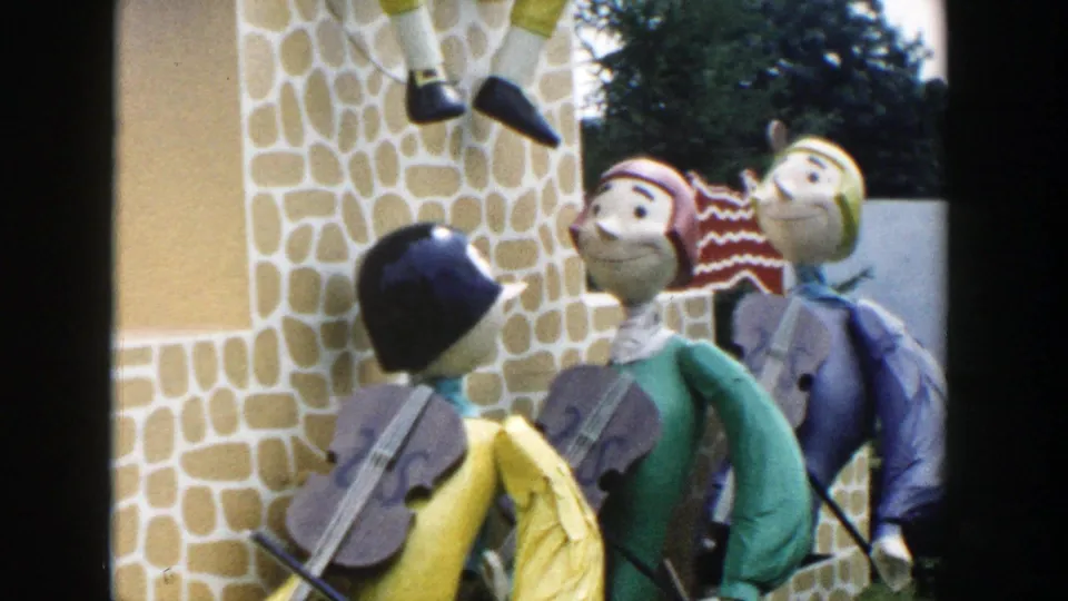 1954 Wisconsin Dells home-movie frame showing colorful decorative figures with violins near a stone-patterned facade.