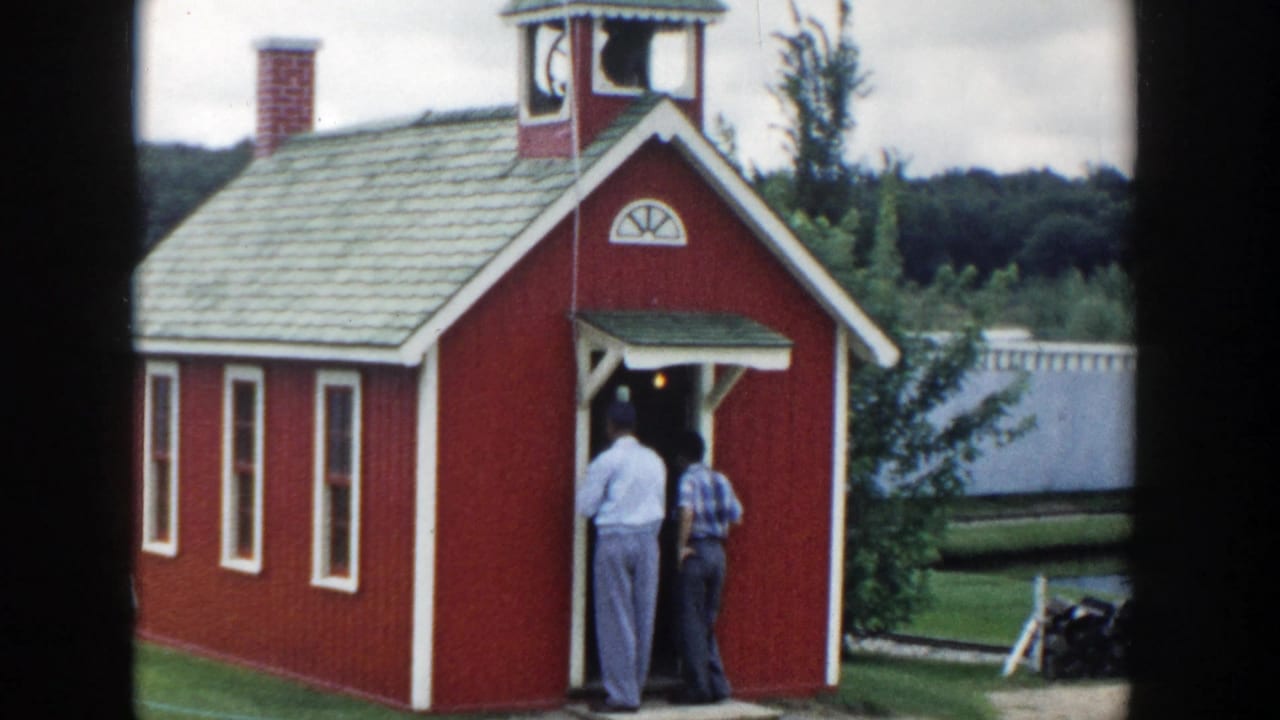 1954 Wisconsin Dells frame showing two adult men at the entrance of a red schoolhouse-style attraction beside a lawn.