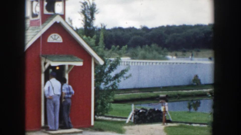 1954 Wisconsin Dells frame showing two men at a small red schoolhouse, with a pond, white fence, and wooded hillside beyond.
