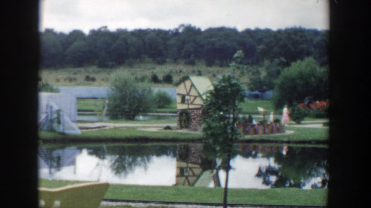 1954 Wisconsin Dells home-movie frame showing a pond, garden path, half-timbered structure, trees, and distant visitors.