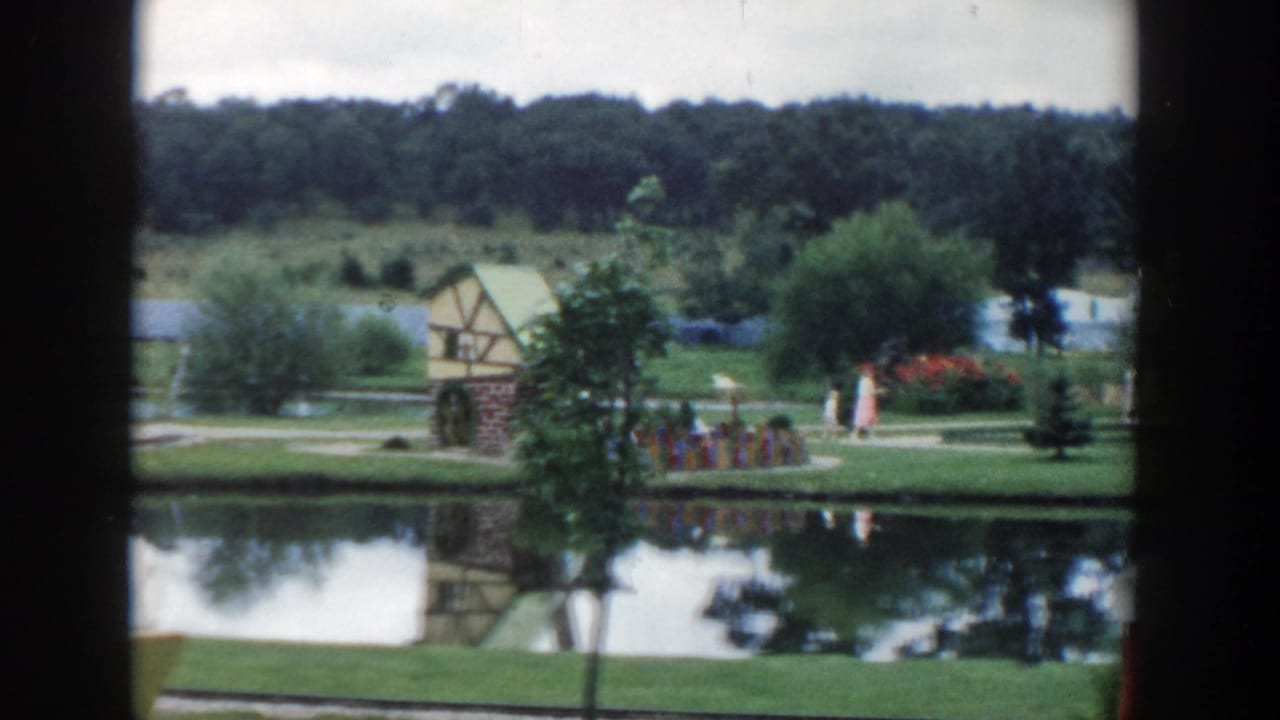1954 Wisconsin Dells frame with a woman and girl walking along a pond path near flowers, trees, and a decorative structure.