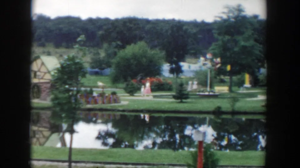 1954 Wisconsin Dells home-movie view of a woman and girl walking beside a pond with flowerbeds, trees, and reflections.