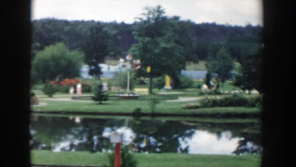 1954 Wisconsin Dells frame showing the woman and girl farther along the pond path, with flowers, trees, and visitors in the background.