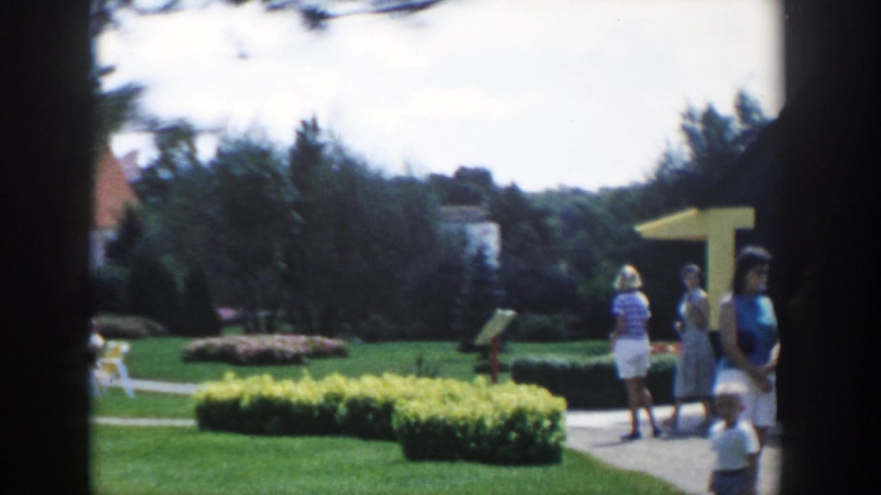 1954 Wisconsin Dells home-movie frame showing women near a landscaped garden path with hedges and a yellow awning.
