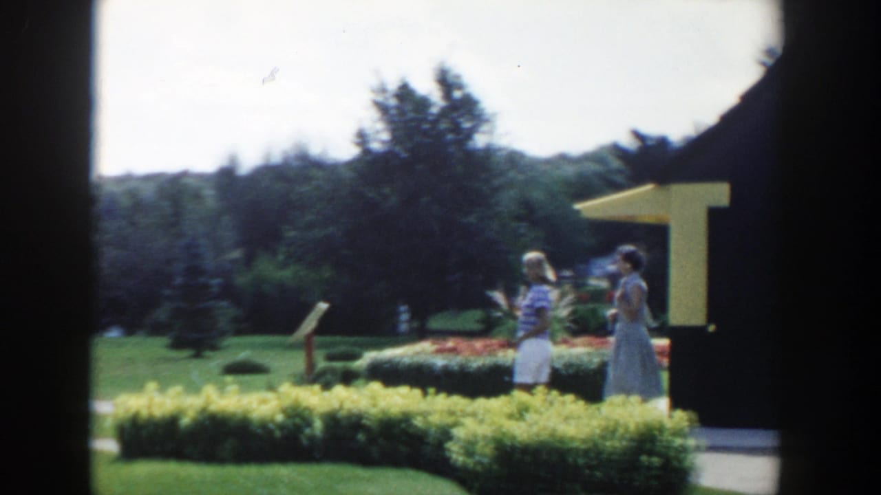 1954 Wisconsin Dells frame of two women conversing in a landscaped garden near a yellow-awning structure.