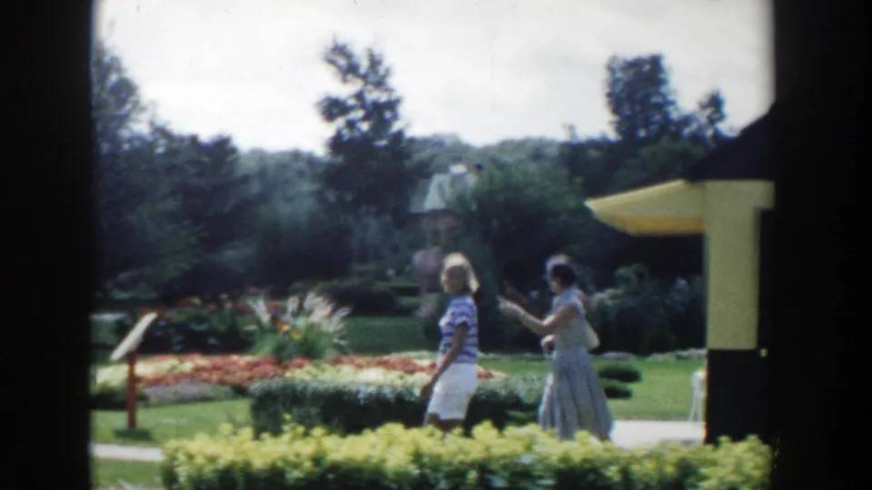 1954 Wisconsin Dells home-movie frame showing two women walking along a garden path past planted beds and hedges.