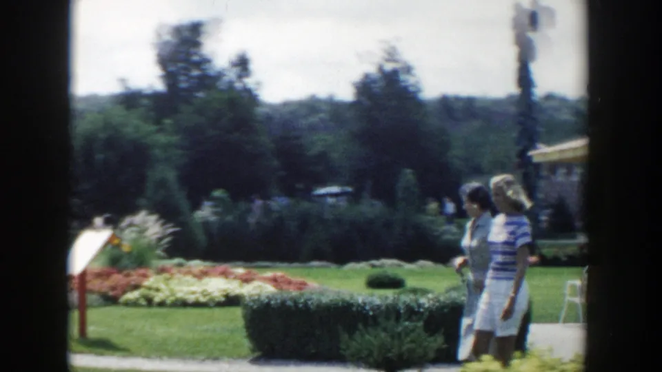 1954 Wisconsin Dells frame showing women walking beside a hedge with flowerbeds, lawns, and trees behind them.