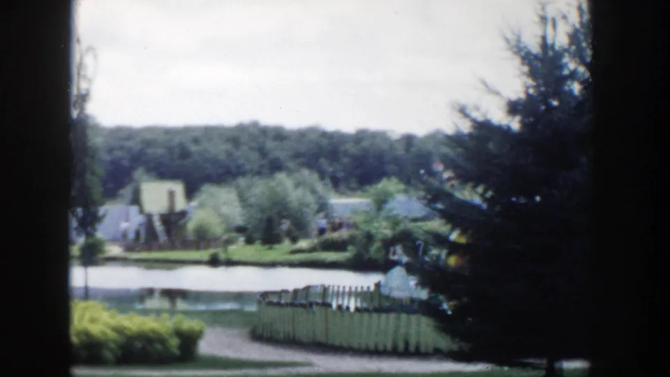 1954 Wisconsin Dells 8mm frame showing a landscaped pond, fencing, trees, path, and distant park visitors.