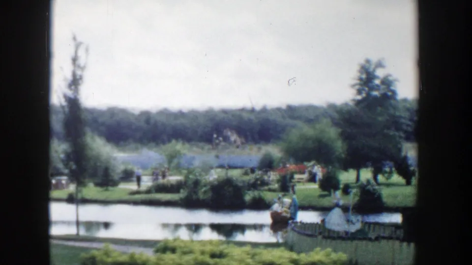 1954 Wisconsin Dells park scene with pond, manicured planting beds, trees, distant visitors, and foreground fencing.