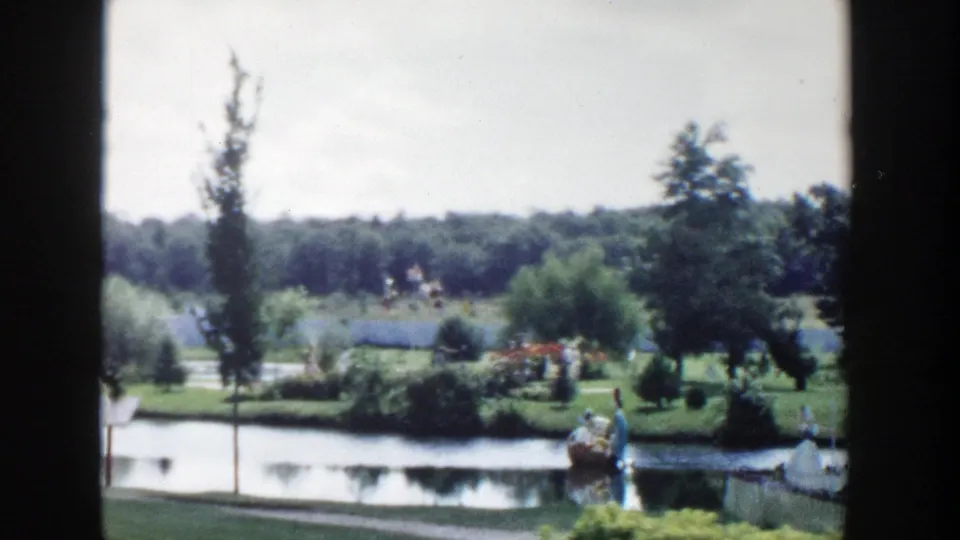 1954 Wisconsin Dells home-movie view of a pond, green lawn, trees, and small distant tourist figures framed by dark film edges.