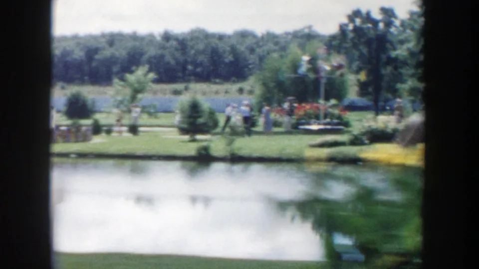 A soft 8mm view of the Wisconsin Dells garden pond shows lawns, shrubs, trees, and small groups of visitors along the far side.