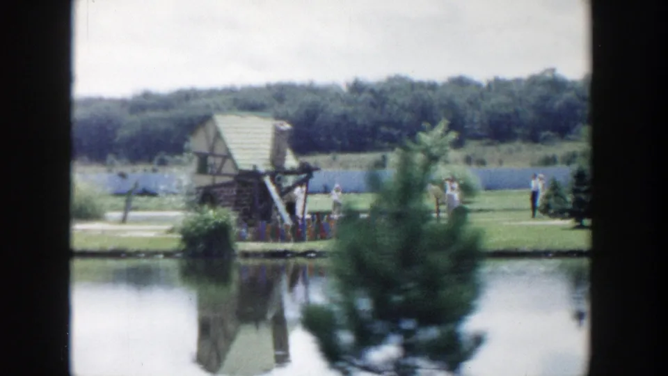 Wisconsin Dells tourist-garden scene with a small gazebo-like structure beside the pond, reflected in the water with visitors nearby.