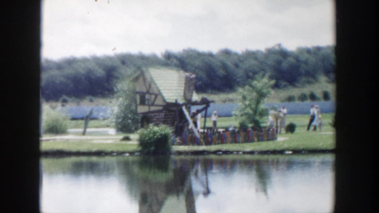 1954 Wisconsin Dells home-movie frame showing a small rustic structure by a pond, with visitors and trees in the background.