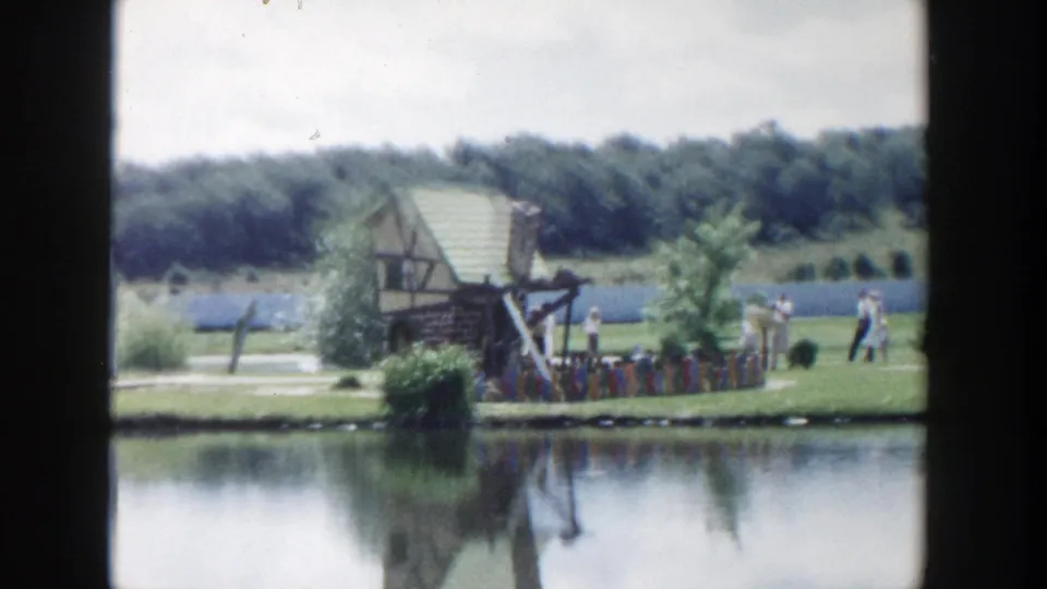 1954 Wisconsin Dells home-movie frame showing a small rustic structure by a pond, with visitors and trees in the background.