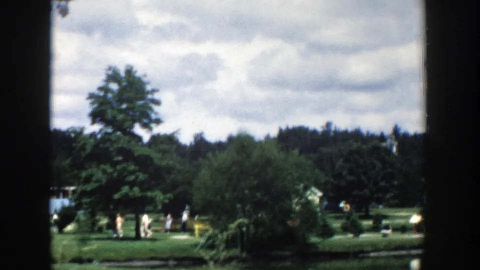 1954 Wisconsin Dells home-movie view of a pond, large trees, lawn, and distant visitors under a cloudy sky.