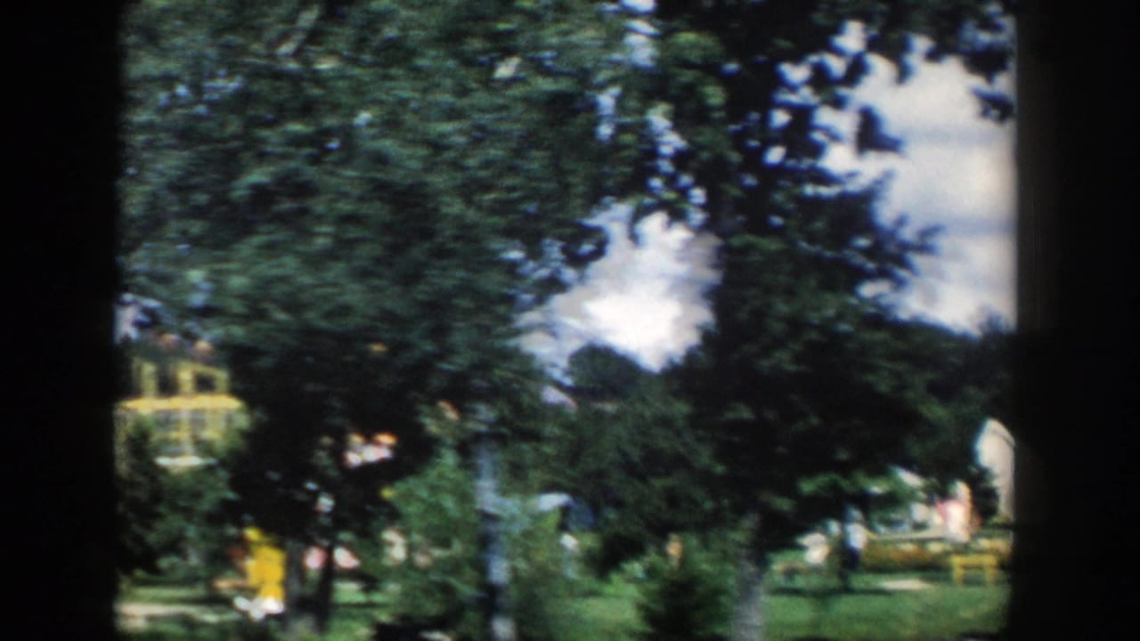 1954 Wisconsin Dells home-movie frame with leafy trees framing bright fantasy-style buildings and paths in a visitor attraction.
