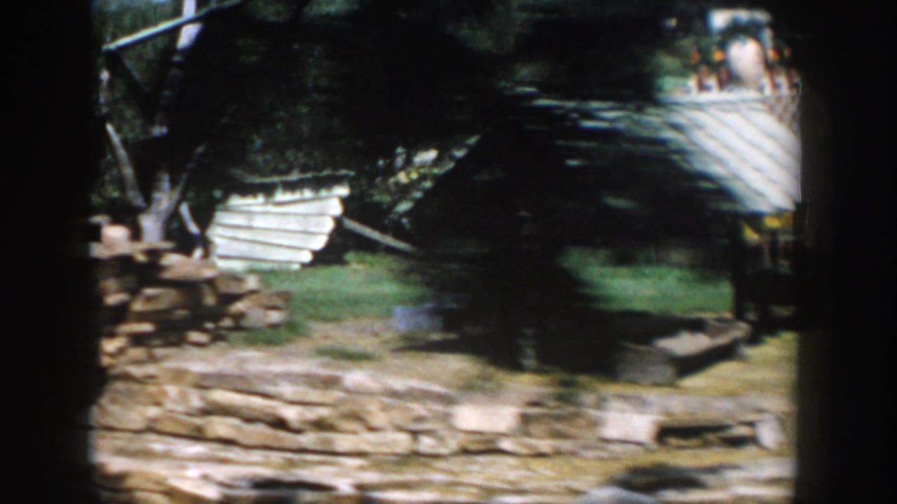 Drive-by view of stone paths, grass, trees, and a small rustic roofed structure.