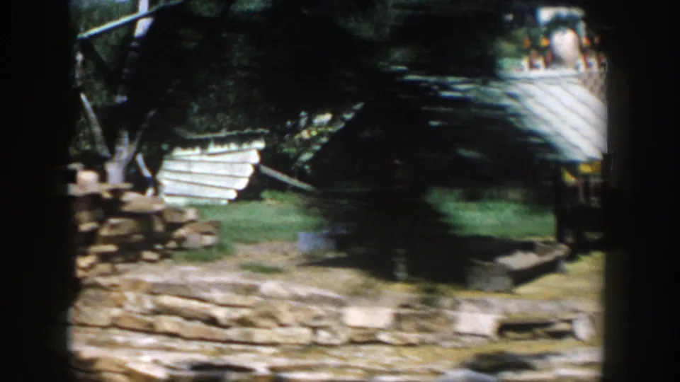 1954 Wisconsin Dells home-movie frame showing stone edging, lawn, trees, and a low rustic shelter in a playground-like area.