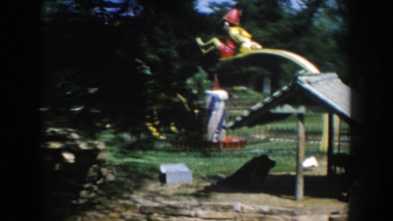 Playground scene with a large curved yellow feature, trees, fencing, and a distant visitor.