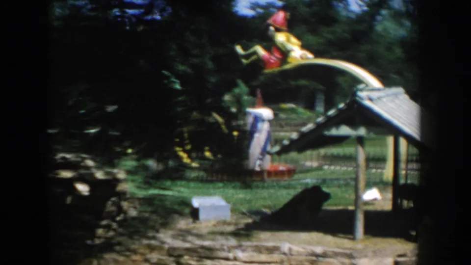 1954 Wisconsin Dells frame with a colorful novelty play sculpture, curved yellow ring, lawn, fence, and a visitor in the background.