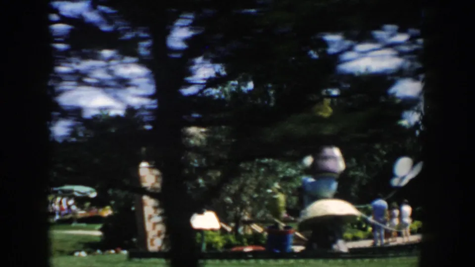 1954 Wisconsin Dells frame showing a large playground figure, shaded trees, stonework, lawn, and several distant visitors.