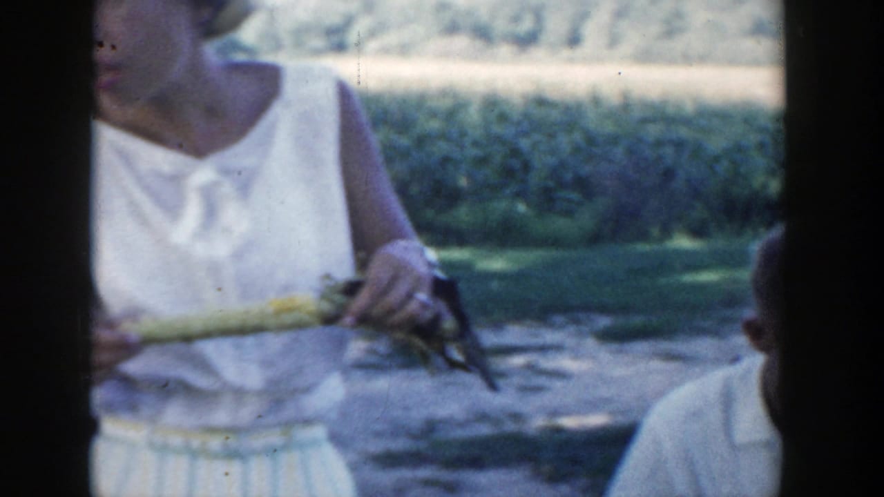 1954 Wisconsin Dells home-movie frame showing a woman in a white sleeveless dress holding corn while seated people gather nearby.