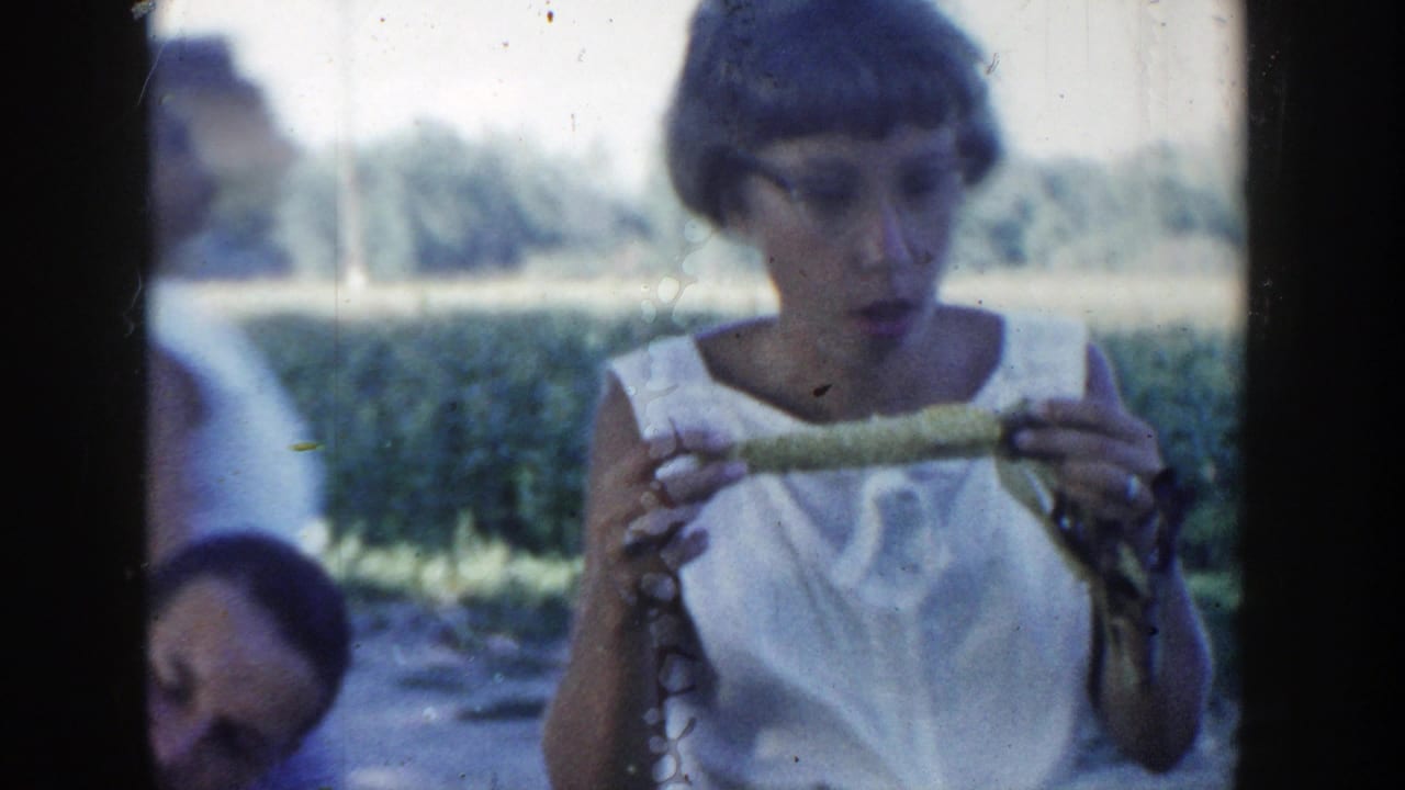 Woman examines or eats corn while people sit behind her outdoors.