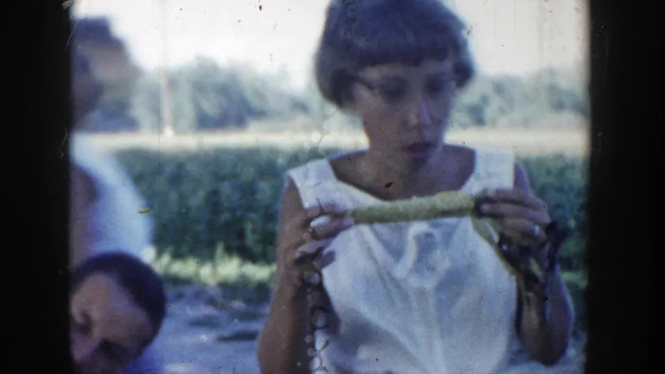 1954 Wisconsin Dells frame with a woman holding corn in both hands, with other adults seated in the shaded background.