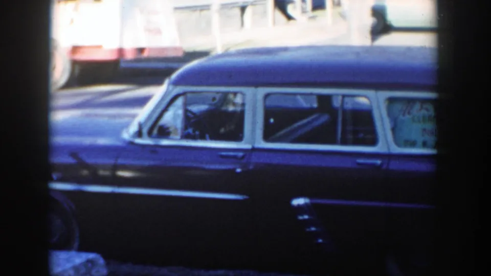 Mid-pan street view shows the parked dark vehicle's side and windows while a larger red-and-white vehicle passes behind it.