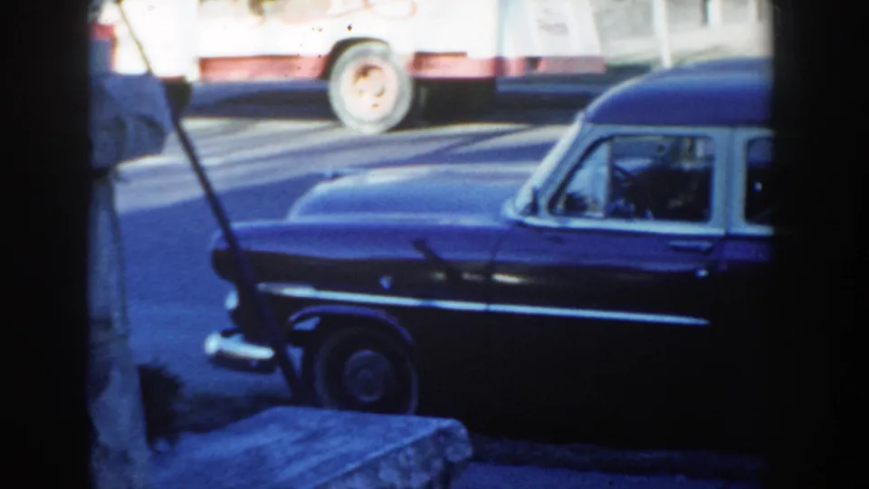 The pan reaches the front half of the dark hearse-style vehicle, with chrome trim, windshield, curb, and passing traffic in view.