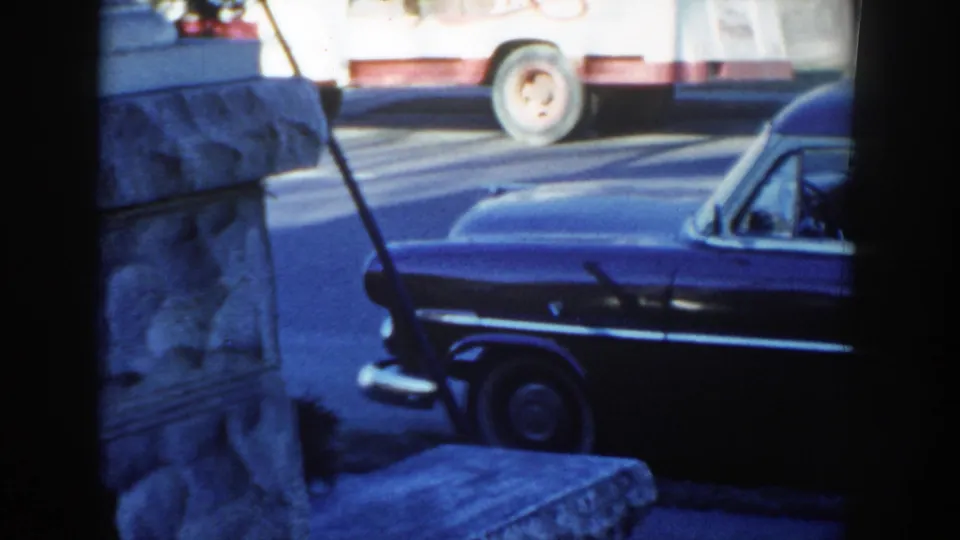 1954 Wisconsin Dells home-movie frame showing part of a dark station wagon parked near a stone wall, with a truck passing or parked above.