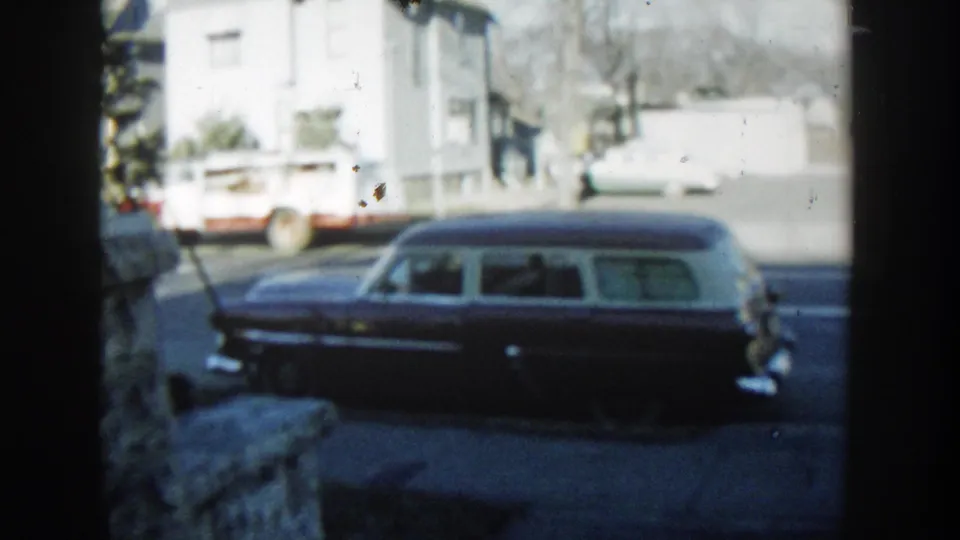 1954 Wisconsin Dells frame showing a dark station wagon with a pale roof parked on a quiet street, with houses and a white truck behind it.