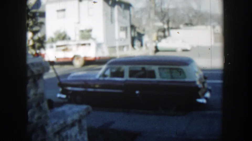 1954 Wisconsin Dells street view with a dark station wagon parked in profile, framed by a stone wall and bright residential background.