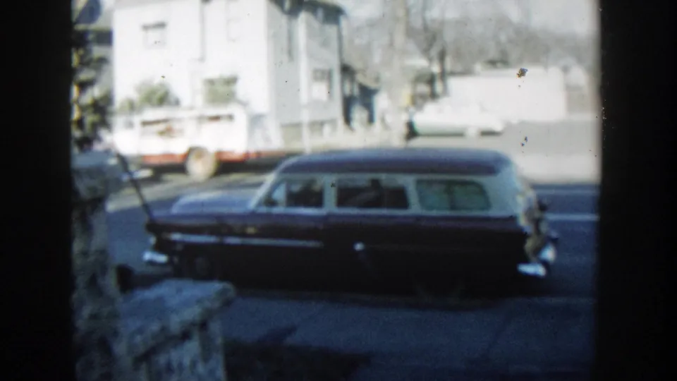 1954 Wisconsin Dells home-movie frame of a dark station wagon parked along the street, with a white truck and light-colored buildings behind it.