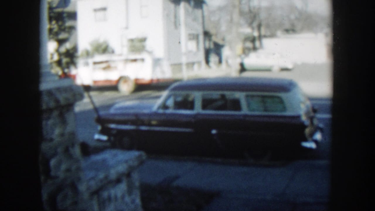 A dark station wagon with a light roof sits on a Wisconsin Dells street, with a utility truck and houses in the background.