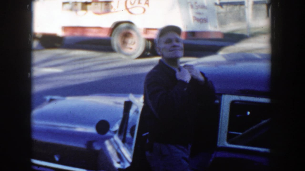 Man adjusts his jacket next to a parked vintage car with a truck visible across the street.