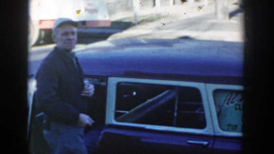 In Wisconsin Dells in 1954, a man wearing a grey cap and dark jacket stands next to the blue-and-white station wagon as traffic passes behind him.