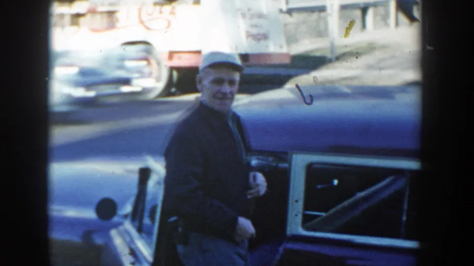 Wisconsin Dells 1954 frame of a smiling man in a grey cap beside the blue station wagon, with passing cars and a Pepsi truck in the background.