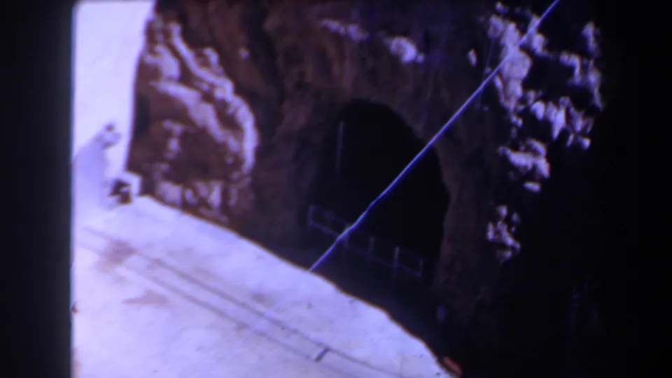 1958 Texas 8mm frame showing a rocky wall, dark cave entrance, pale ground, and a small fence or rail near the opening.