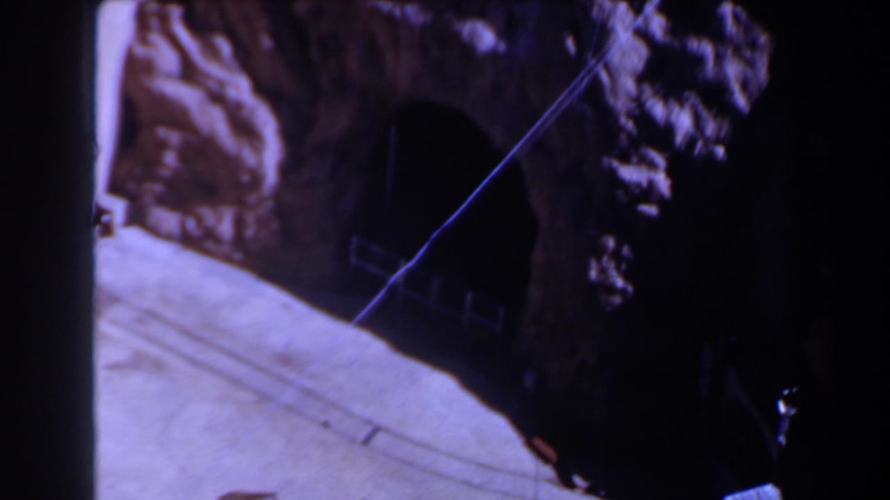 1958 Texas 8mm frame showing a dark tunnel opening cut into rugged rock, with a pale concrete ramp or ledge and a diagonal wire across the view.