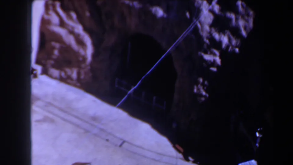 1958 Texas 8mm frame showing a dark tunnel opening cut into rugged rock, with a pale concrete ramp or ledge and a diagonal wire across the view.